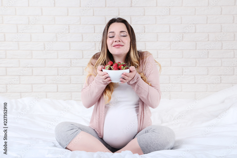 Beautiful pregnant woman with strawberries in bowl sitting on bed Beautiful pregnant woman with strawberries in bowl sitting on bed