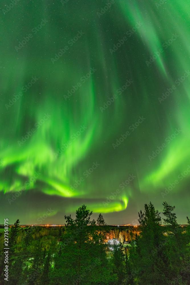 View from scandinavian hill to bright green aurora lights almost on the whole sky over tree tops in Sweden, river, clear skies with a lot of stars, pine trees and autumn colored leaves on trees