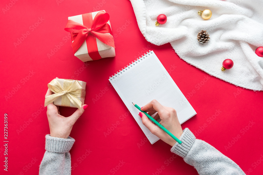 Female hands, girl holds a gift and writes in Notepad, red table with gift boxes for Christmas and New year, green pensil, background with copy space, for advertisement, top view