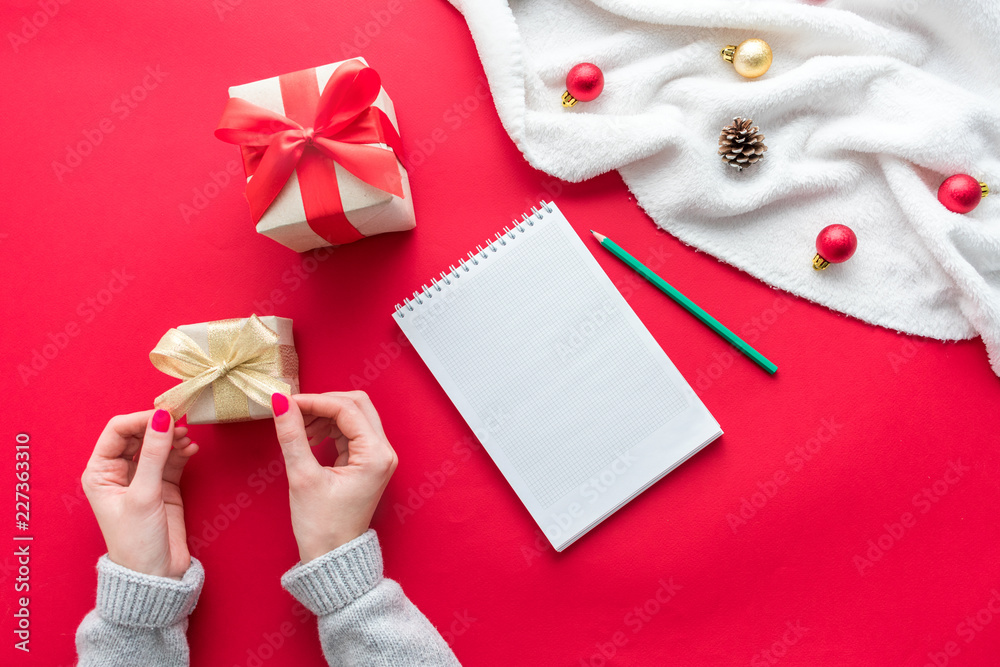 Female hands,  woman holding a box with a gift, white Notepad, red table with gift boxes for Christmas and New year, green pensil, background with copy space, for advertisement, top view