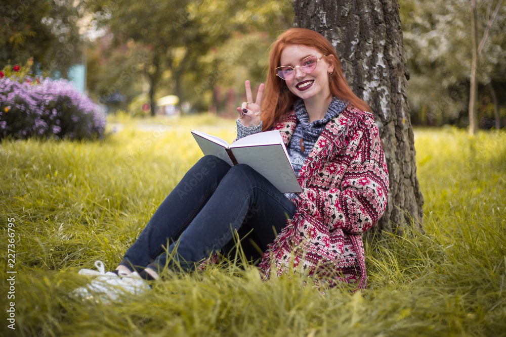 Cheerful woman with freckles and dark lipstick wearing sunglasses reading book outdoors in park sit under big tree on grass with flowers on the background showing peace gesture. Cheerful woman with freckles and dark lipstick wearing sunglasses reading book outdoors in park sit under big tree on grass with flowers on the background showing peace gesture.