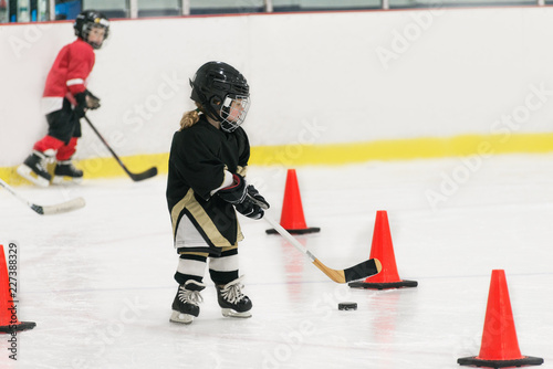 A little cute hockey girl is training on ice. Girl is wearing in full hockey equipment: helmet, gloves, skates. stick, puck. She is skating among orange cones. Blurred young boy is on the background.