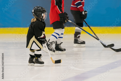 The little cute hockey girl is on the ice wearing in full equipment: hockey helmet, gloves, stick, skates. Figures of two teenagers hockey players are on the background.
