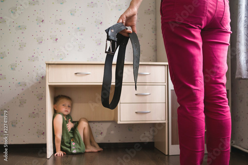 Domestic violence. Mother holding the belt in her hand to punish her small daughter. Scared child hiding under a table and sitting there terrified by physical punishment