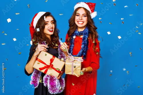 nice portrait of two happy young women in santa claus hat with gift .Christmas concept. in evening dresses on party over blue background. firecrackers in the background.