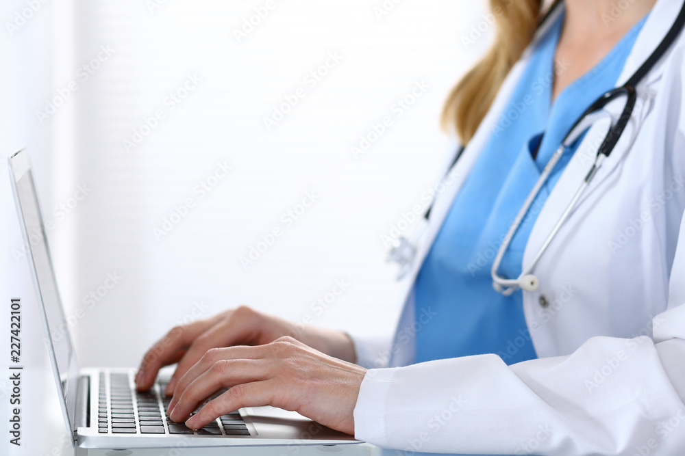 Doctor typing on laptop computer while sitting at the glass desk in hospital office. Physician at work. Medicine and healthcare concept Doctor typing on laptop computer while sitting at the glass desk in hospital office. Physician at work. Medicine and healthcare concept