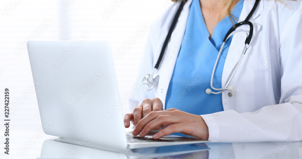 Doctor typing on laptop computer while sitting at the glass desk in hospital office. Physician at work. Medicine and healthcare concept Doctor typing on laptop computer while sitting at the glass desk in hospital office. Physician at work. Medicine and healthcare concept