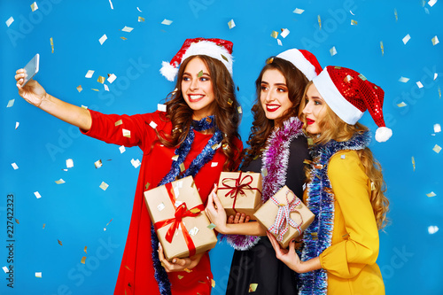 portrait of three happy young women in santa claus hat with gift make selfie .Christmas concept. in evening dresses on party over blue background. firecrackers in the background