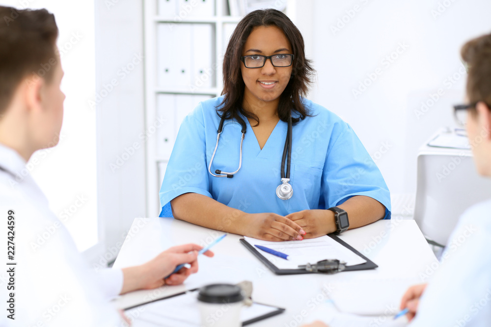 Female african american medical doctor with colleagues in background at hospital. Medicine and health care concept Female african american medical doctor with colleagues in background at hospital. Medicine and health care concept