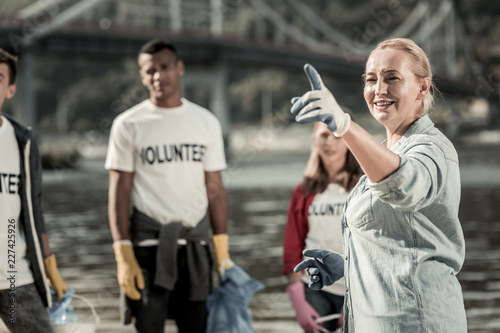 Woman with volunteers. Mature beaming blonde-haired woman wearing blue gloves talking to young volunteers standing near the beach