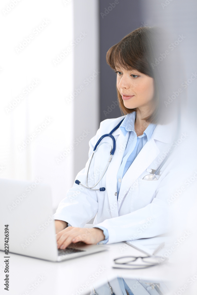 Doctor typing on laptop computer while sitting at the white table in hospital office. Physician at work. Medicine and healthcare concept Doctor typing on laptop computer while sitting at the white table in hospital office. Physician at work. Medicine and healthcare concept