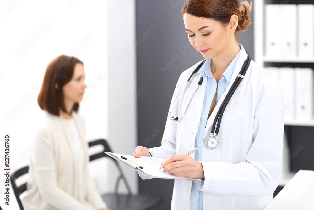 Doctor woman at work. Portrait of female physician filling up medical form while standing near reception desk at clinic or emergency hospital. Patient woman sitting at the background. Medicine concept Doctor woman at work. Portrait of female physician filling up medical form while standing near reception desk at clinic or emergency hospital. Patient woman sitting at the background. Medicine concept