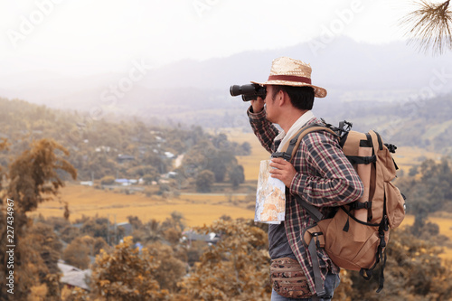 Young man with backpack and holding a binoculars and map paper is standing top of a mountain.