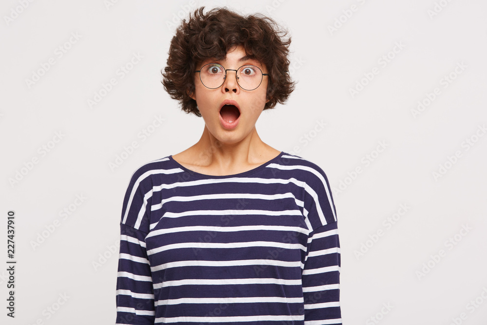 Studio shot of young beautiful girl looks amaized with open mouth, wears white and blue stripped sweatshirt and transparent black-framed round glasses isolated over white background Studio shot of young beautiful girl looks amaized with open mouth, wears white and blue stripped sweatshirt and transparent black-framed round glasses isolated over white background