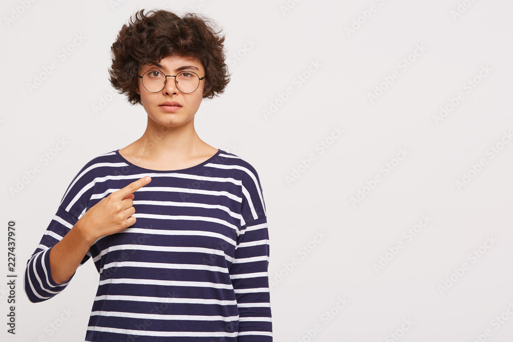 young beautiful girl looks dissatisfied, one eyebrow raised points to the side with finger, wears white and blue stripped sweatshirt and transparent black-framed round glasses isolated young beautiful girl looks dissatisfied, one eyebrow raised points to the side with finger, wears white and blue stripped sweatshirt and transparent black-framed round glasses isolated