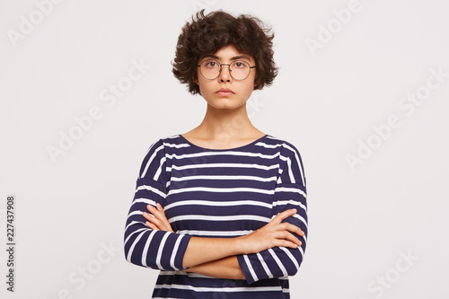 Closeup of calm serious emotionless young curly brownhair woman wears white and blue stripped sweatshirt and transparent black-framed round glasses keeps arms crossed isolated over white background photo