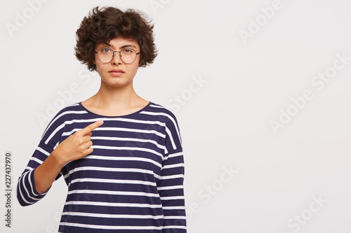 young beautiful girl looks dissatisfied, one eyebrow raised points to the side with finger, wears white and blue stripped sweatshirt and transparent black-framed round glasses isolated 