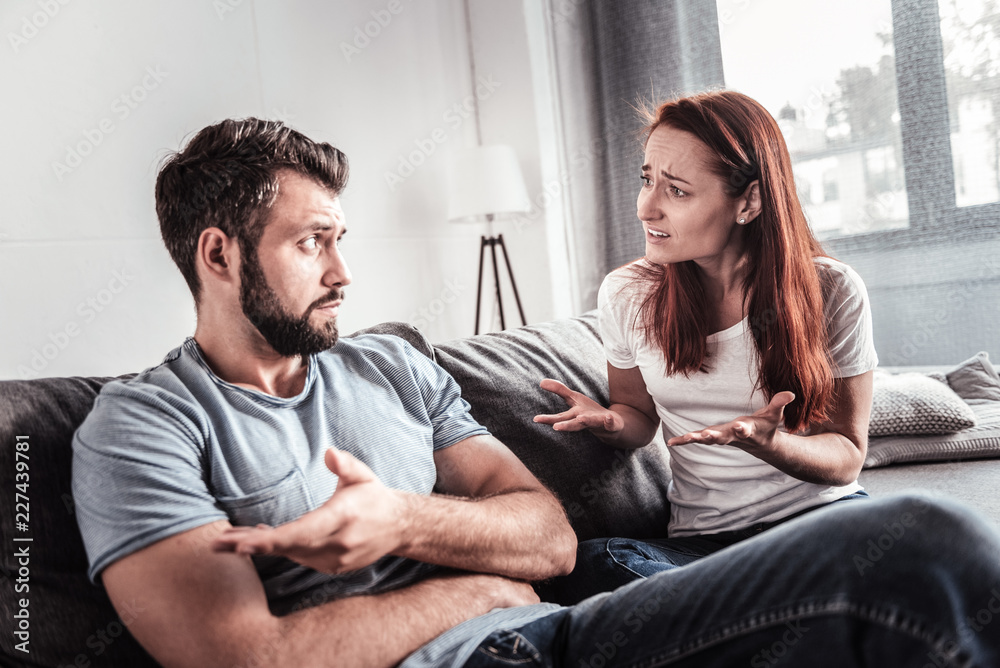 Problems in family. Unhappy cheerless young couple sitting together and talking to each other while having quarrel Problems in family. Unhappy cheerless young couple sitting together and talking to each other while having quarrel