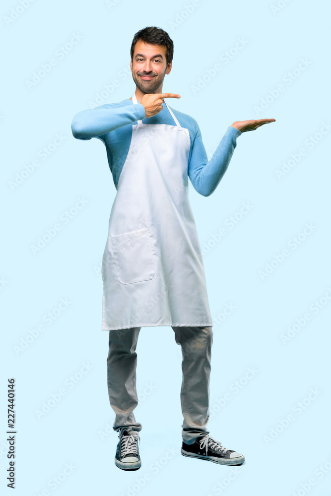 Full body of Man wearing an apron holding copyspace imaginary on the palm to insert an ad on blue background