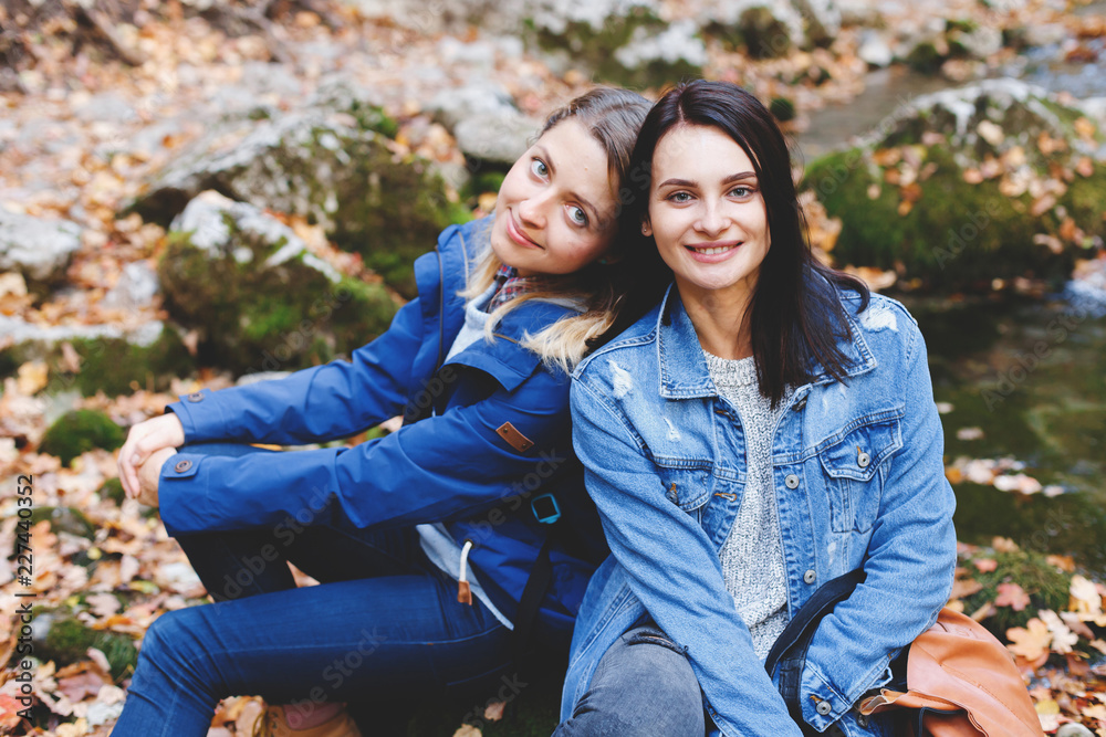 Two young girls friends sitting on a rock near the river in the autumn forest. Walk in the woods. Girlsfriends hiking at fall. Two young girls friends sitting on a rock near the river in the autumn forest. Walk in the woods. Girlsfriends hiking at fall.