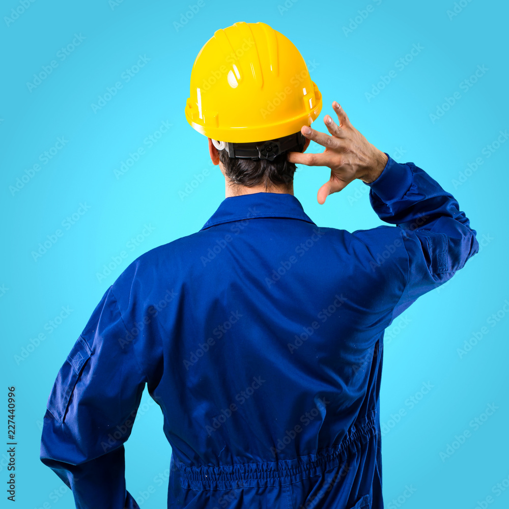 Young workman with helmet on back position looking back while scratching head on blue background Young workman with helmet on back position looking back while scratching head on blue background