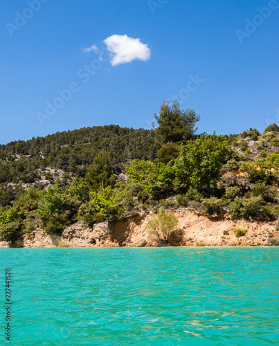Beautiful view of St. Croix lake in Verdon and the forested hills of the coastline. (Provence, France) Sunlight on the water surface. Blue sky with lonely white cloud.