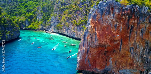 Multiple Boats in Loh Samah Bay, Thailand - Tropical Cove Surrounded by Lush Island Cliff Face With Turquoise Blue Water and Visible Coastal Reef - Aerial Overhead View