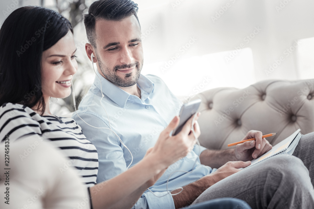 Music lovers. Attractive brunette female feeling happiness while looking at telephone, sitting near her partner Music lovers. Attractive brunette female feeling happiness while looking at telephone, sitting near her partner