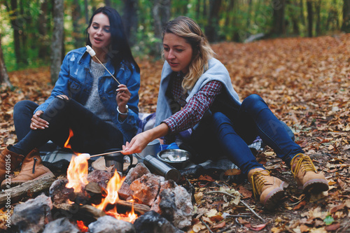 Two young girls friends roasting sweet marshmallow on a fire in the evening in the autumn forest.