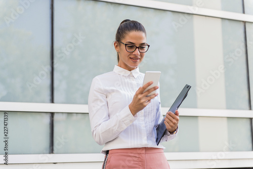 Successful young businesswoman using smartphone and holding documents outdoors.
