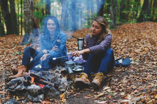 Two young girls girlfriends roasting sweet marshmallow on a fire in the evening in the autumn forest.