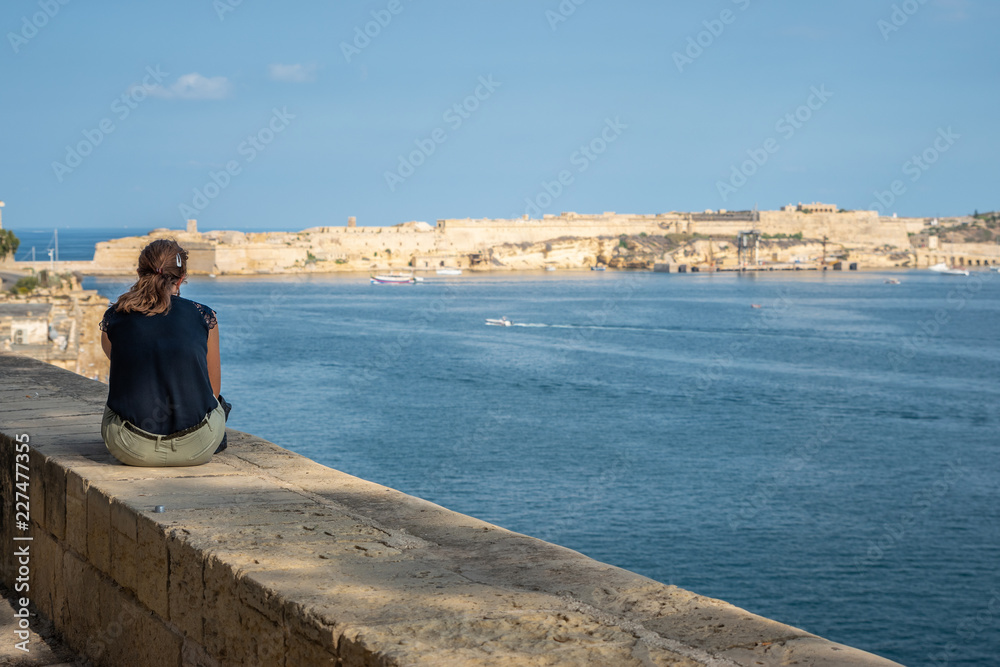 Wide angle view of a young lady sitting on the harbour wall looking looking at the view. Horizontal. Malta Grand hourbour
