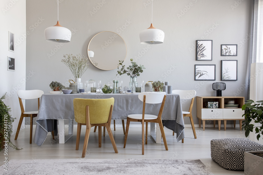 Pouf, plants and round mirror in bright dining room interior with chairs at table under lamps. Real photo - obrazy, fototapety, plakaty 