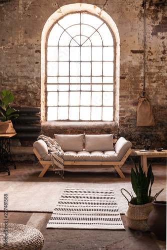 Window above grey wooden sofa in spacious loft interior in wabi sabi style with plant and carpet. Real photo