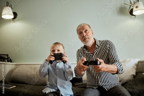 grandfather and grandson playing video game with joysticks in bed room while sitting on couch photo