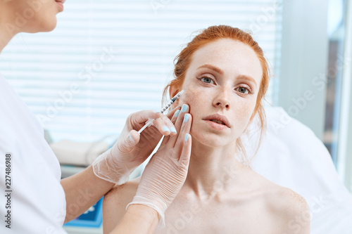 Red haired woman having facial mesotherapy in beauty salon, adult woman making facial injections for improving state of the skin and for the revitalisation and regeneration thereof. photo