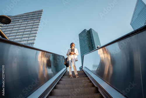 Stylish businesswoman in white suit going down on the escalator at the business centre outdoors with skyscrapers on the background in Paris