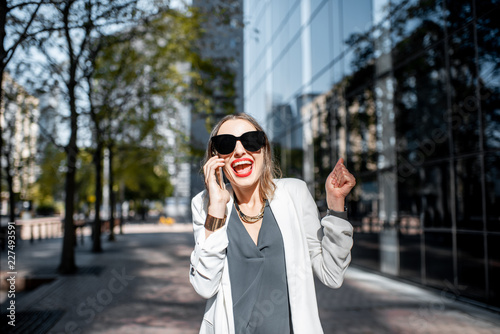 Excited business woman in white suit talking with smartphone at the financial district with modern buildings on the background