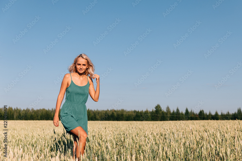 Young girl blonde in a green dress walking on a yellow field with ears of corn near the forest Young girl blonde in a green dress walking on a yellow field with ears of corn near the forest