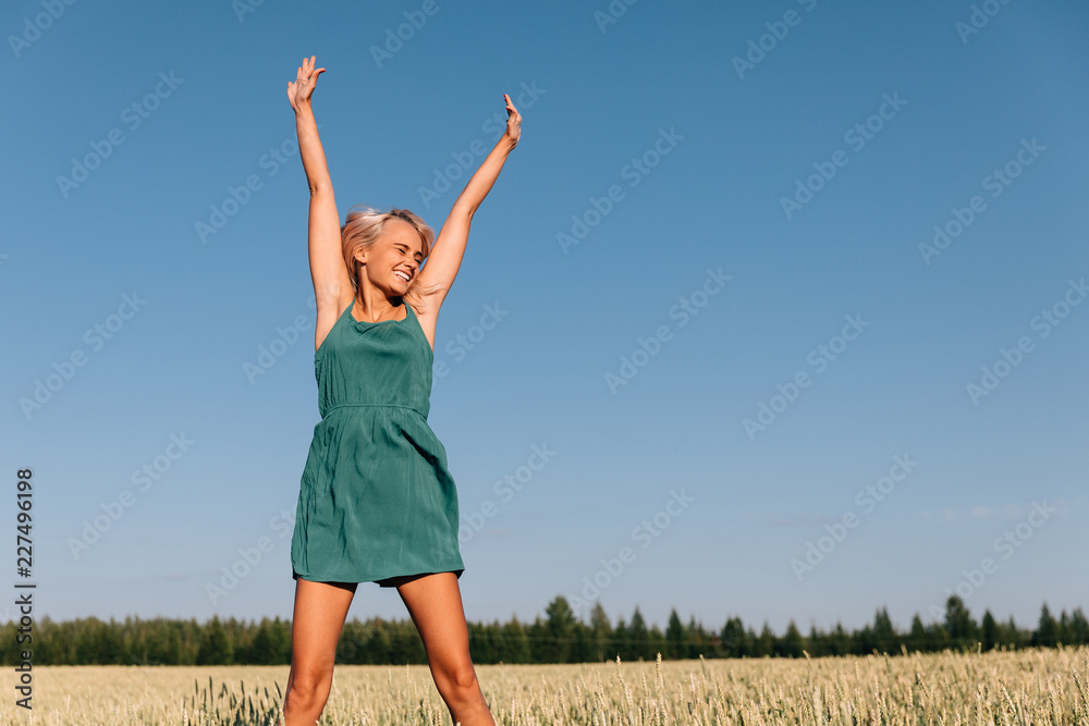 A young girl blonde in a green dress jumps high on the field with ears near the forest A young girl blonde in a green dress jumps high on the field with ears near the forest