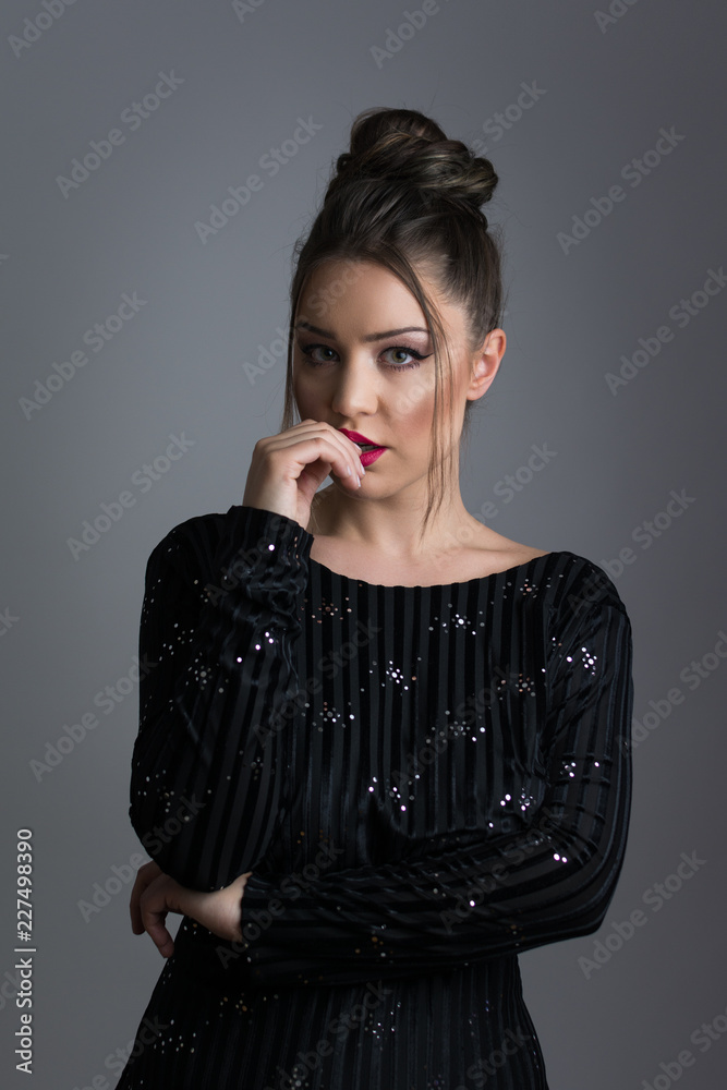 Fototapeta premium Beautiful elegant young woman in shiny black dress, posing, biting her fingers. Studio lighting, dark gray background.
