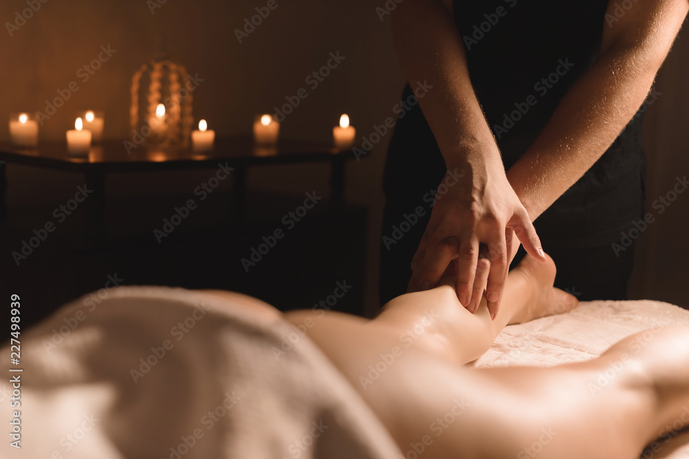 Fototapeta premium Close-up of male hands doing calf massage of female legs in a dark room with candles in the background. Cosmetology and spa treatments