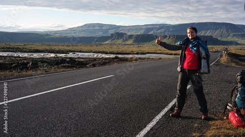 A young woman standing and hitch-hiking by road on Iceland, Icelandic nature with mountains in the background