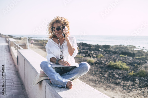 Cheerful beautiful middle age caucasian woman with curly hair smile and speak on the mobile phone sit down near the ocean in technology outdoor activity. yoga crossed legs and laptop for modern people