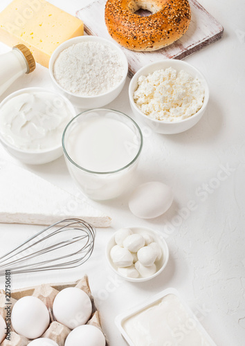 Fresh dairy products on white table background. Glass of milk, bowl of flour and cottage cheese and eggs. Fresh baked bagel with knife. Steel whisk. Top view