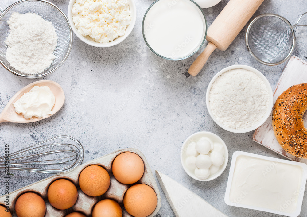 Fresh dairy products on white table background. Glass of milk, bowl of sour cream and cottage cheese and eggs. Fresh baked bagel. Steel whisk. Top view.Space for text