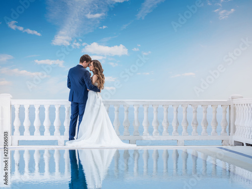 Wedding couple hugging and standing with her back outdoors near the pool. Happy bride and groom in love on their honeymoon. Woman in an elegant dress a man in the suit.