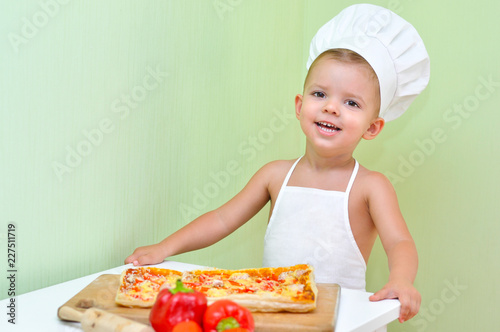 A little boy baker and pizza maker in a white chef's cap and apron is smiling because he cooked delicious and beautiful puff pastry pizza. 