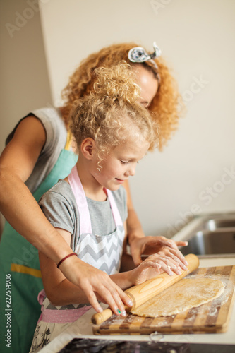 Obraz na plátně Young little girl, Mommy s helper assisting mother in preparing Eastern bread for the holiday, rolling out the dough with mother s hands nearby