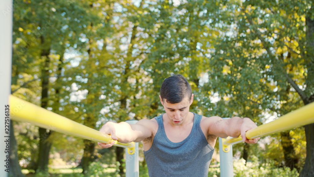 Obraz premium Male athlete performing difficult exercise on gymnastic parallel bars. Slim athlete a very fit guy fitness instructor or a personal trainer working out his arm muscles on outdoor beach gym as part of